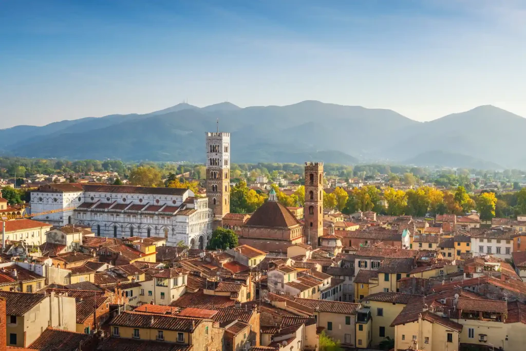 Vista aerea sui tetti in terracotta, i campanili e le chiese romaniche di Lucca con le colline toscane sullo sfondo.