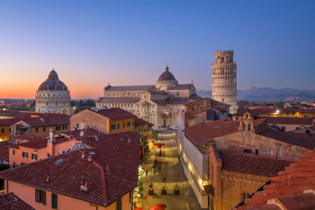 Vista panoramica al tramonto di Piazza dei Miracoli a Pisa con il Battistero, il Duomo e la Torre Pendente illuminata.